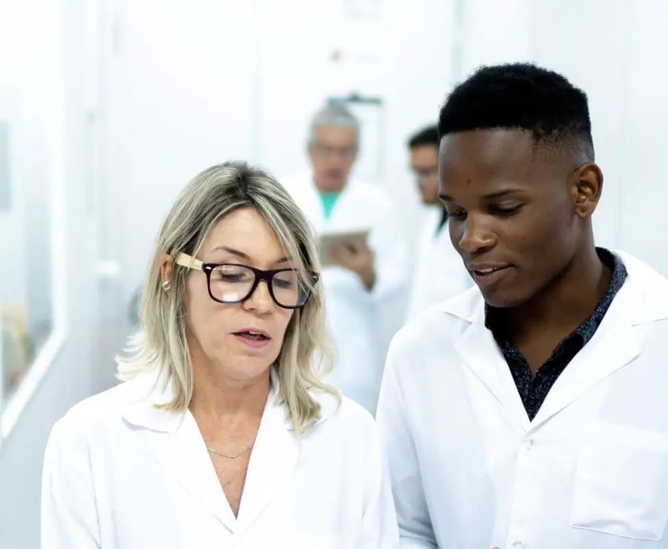 Older white female scientist walking down hallway discussing procedures with young black male scientist.